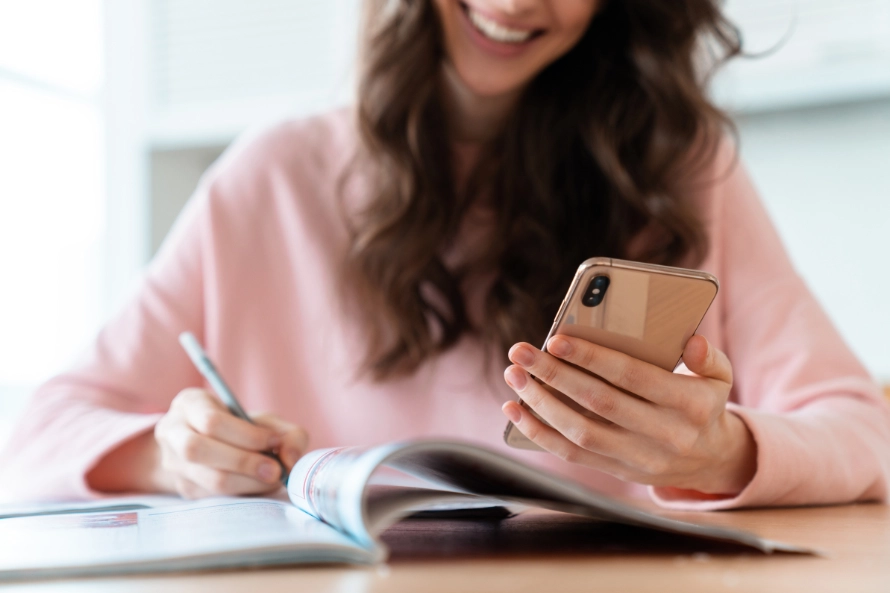 Una mujer joven sentada escribiendo en un cuaderno algo que ve en su celular